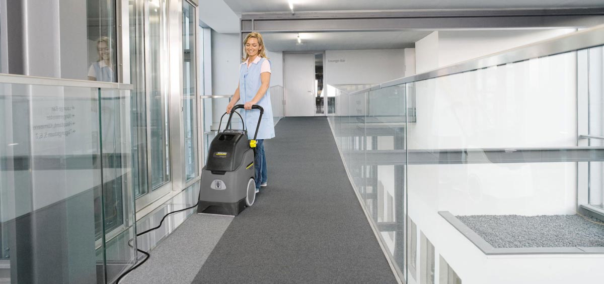 Profesjonalne, kompaktowe urządzenie do czyszczenia zasadniczego BRC 30/15 C firmy Karcher. Kobieta czyszcząca ekstrakcyjnie wykładzinę podłogową na korytarzu w budynku biurowym.